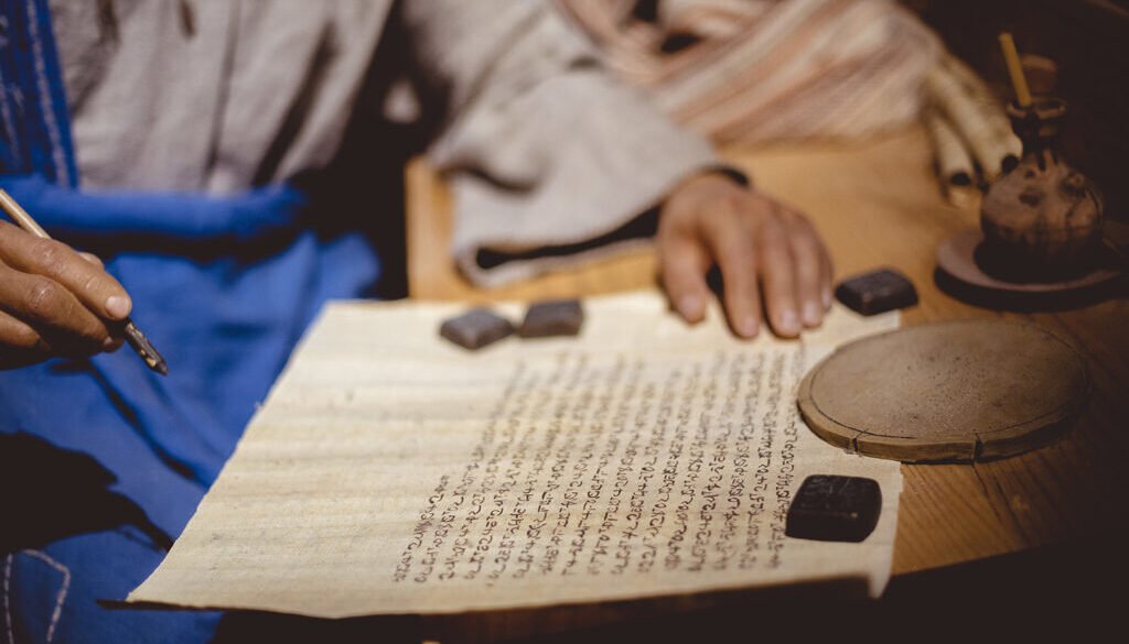 Closeup of a person writing in Hebrew on an aged paper on the table under the lights