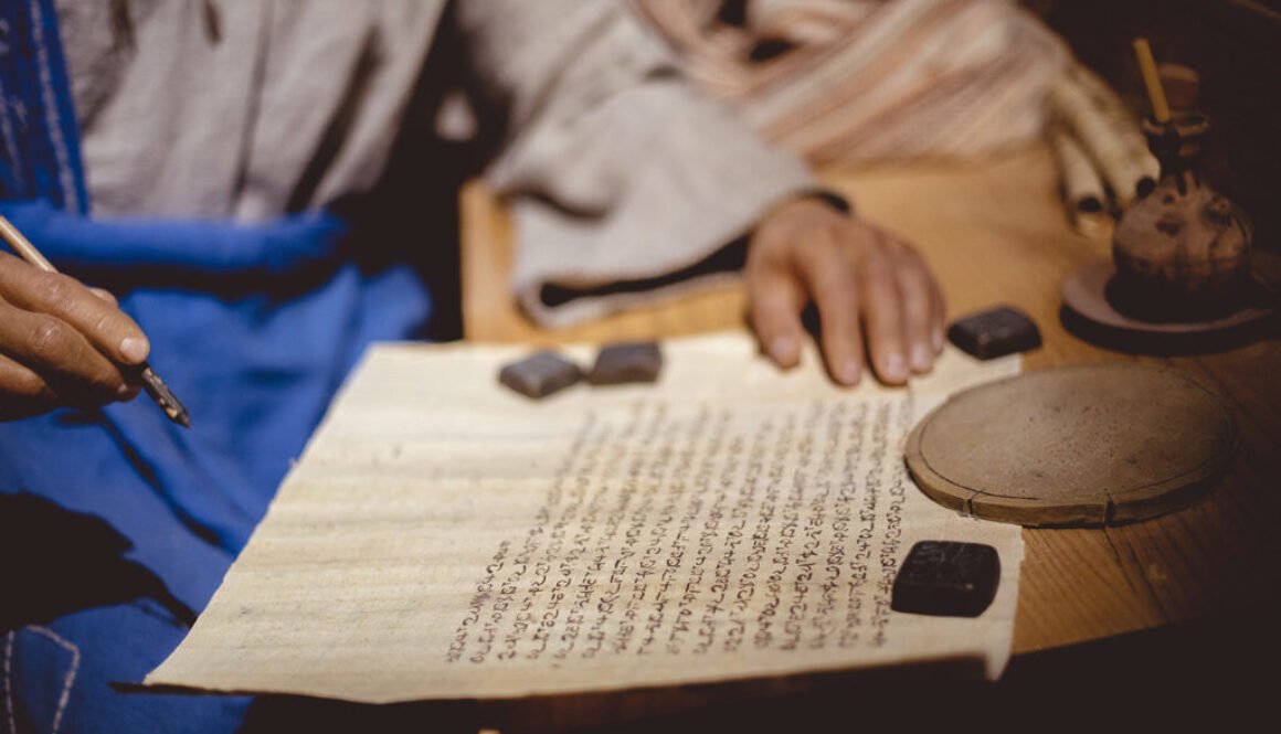 Closeup of a person writing in Hebrew on an aged paper on the table under the lights